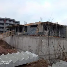 Budowa domu z betonu komórkowego na etapie wznoszenia ścian, widoczne betonowe schody i budynek wielorodzinny w tle.