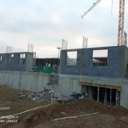Wznoszenie ścian z bloczków betonowych na fundamencie z betonu, widoczny żuraw budowlany w tle oraz składowane materiały budowlane.