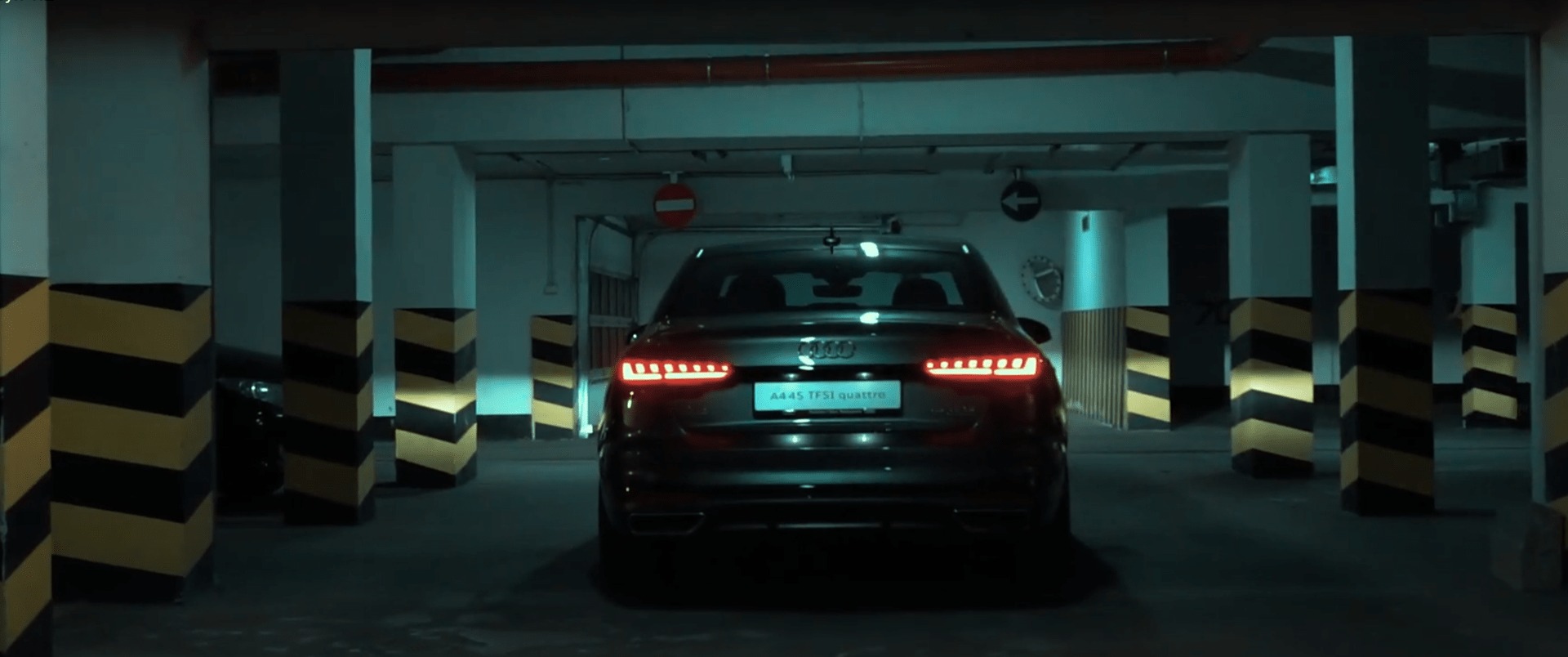 Ciemny samochód Audi A45 TFSI quattro z włączonymi światłami stopu, widziany od tyłu, zaparkowany w garażu podziemnym z kolumnami w żółto-czarne pasy.