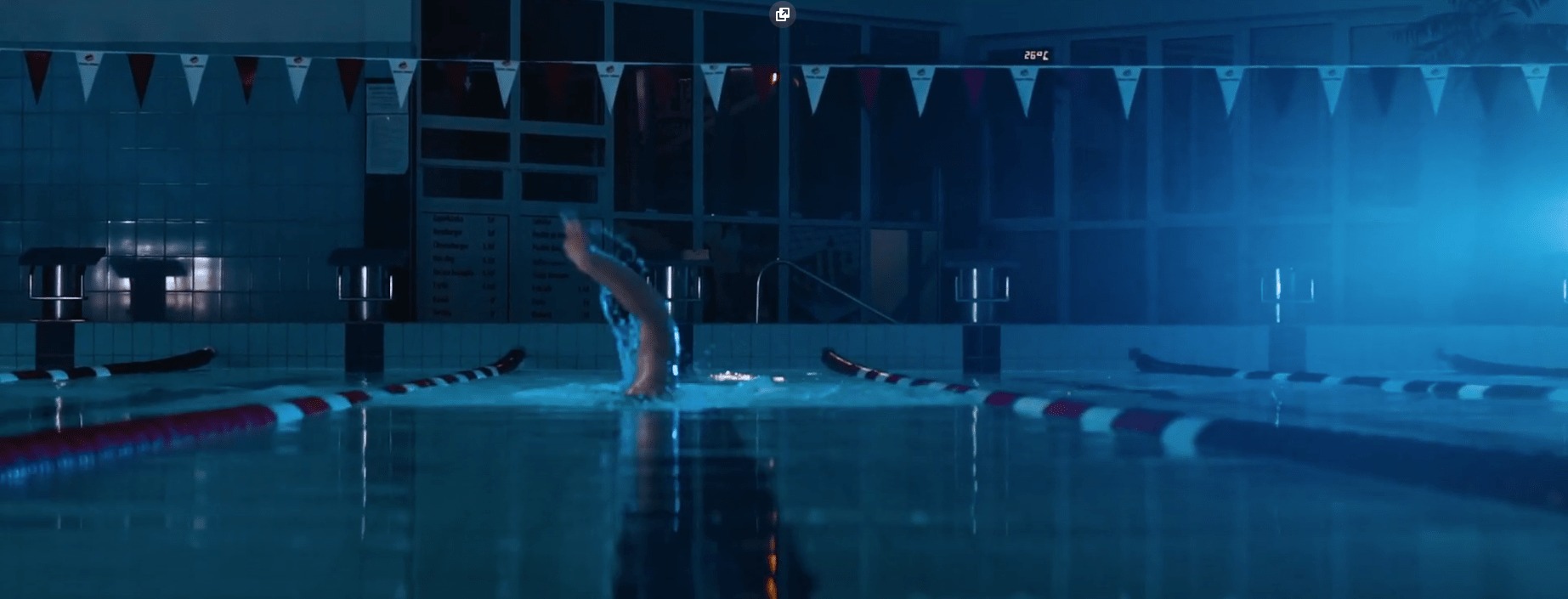 Pływak skaczący do basenu sportowego z oświetleniem LED, widoczne tory pływackie i dekoracyjne flagi.