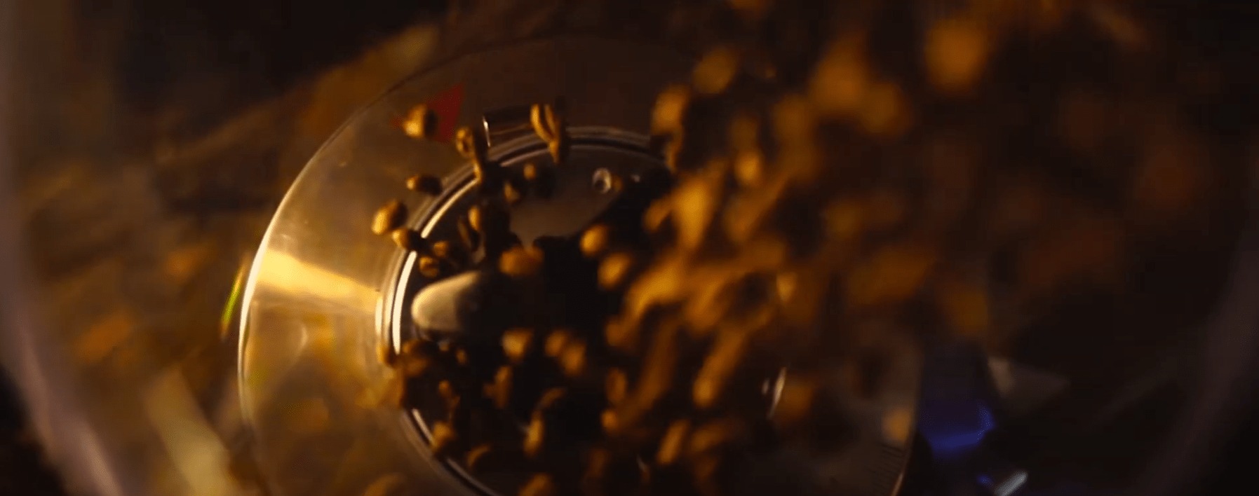 Dynamiczne ujęcie z bliska: złote ziarna w ruchu wewnątrz metalowej maszyny z widocznym mechanizmem obrotowym, prawdopodobnie podczas procesu prażenia. Rozmycie ruchu podkreśla akcję.