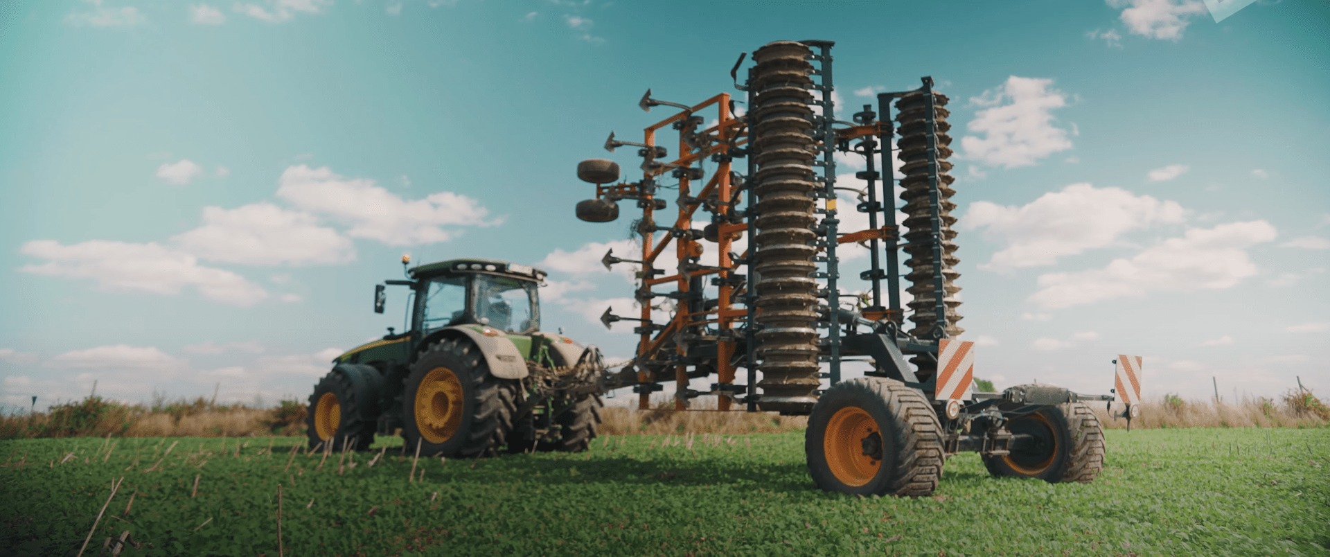 Zielony traktor z pomarańczowymi felgami ciągnie za sobą pomarańczowo-czarne brony talerzowe na tle błękitnego nieba z chmurami.