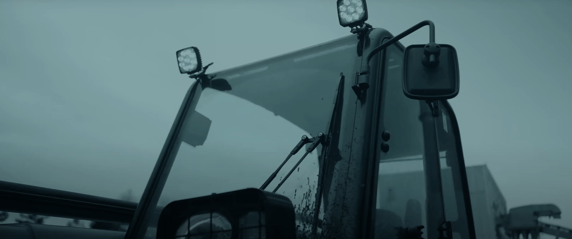 Kabina maszyny budowlanej z reflektorami LED i widocznymi wycieraczkami, w tle pochmurne niebo.