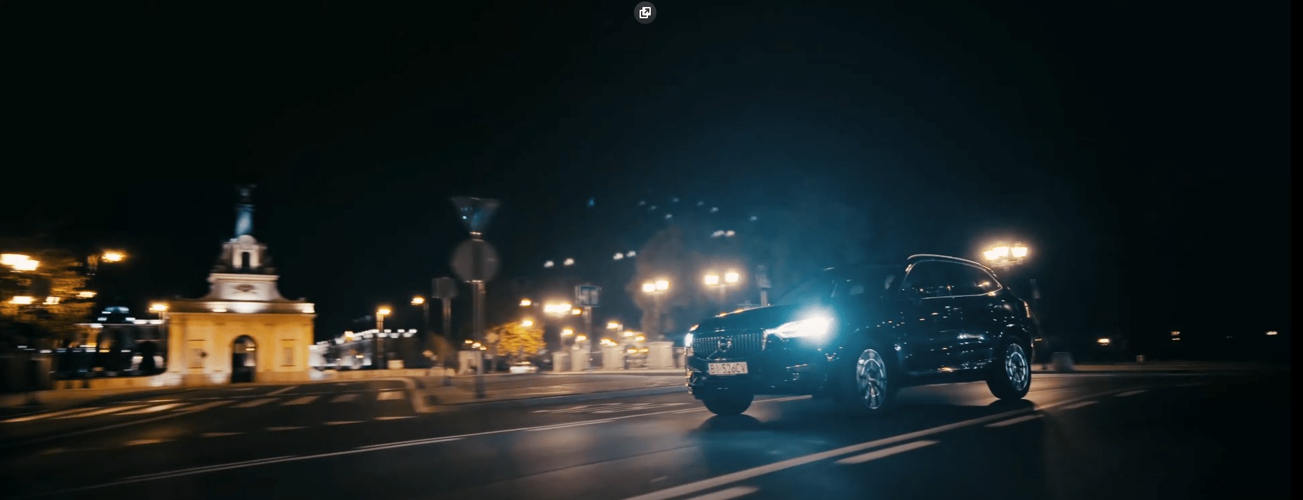 Czarny samochód Volvo SUV z włączonymi światłami mija zabytkową bramę miejską nocą, widoczne latarnie i znaki drogowe.