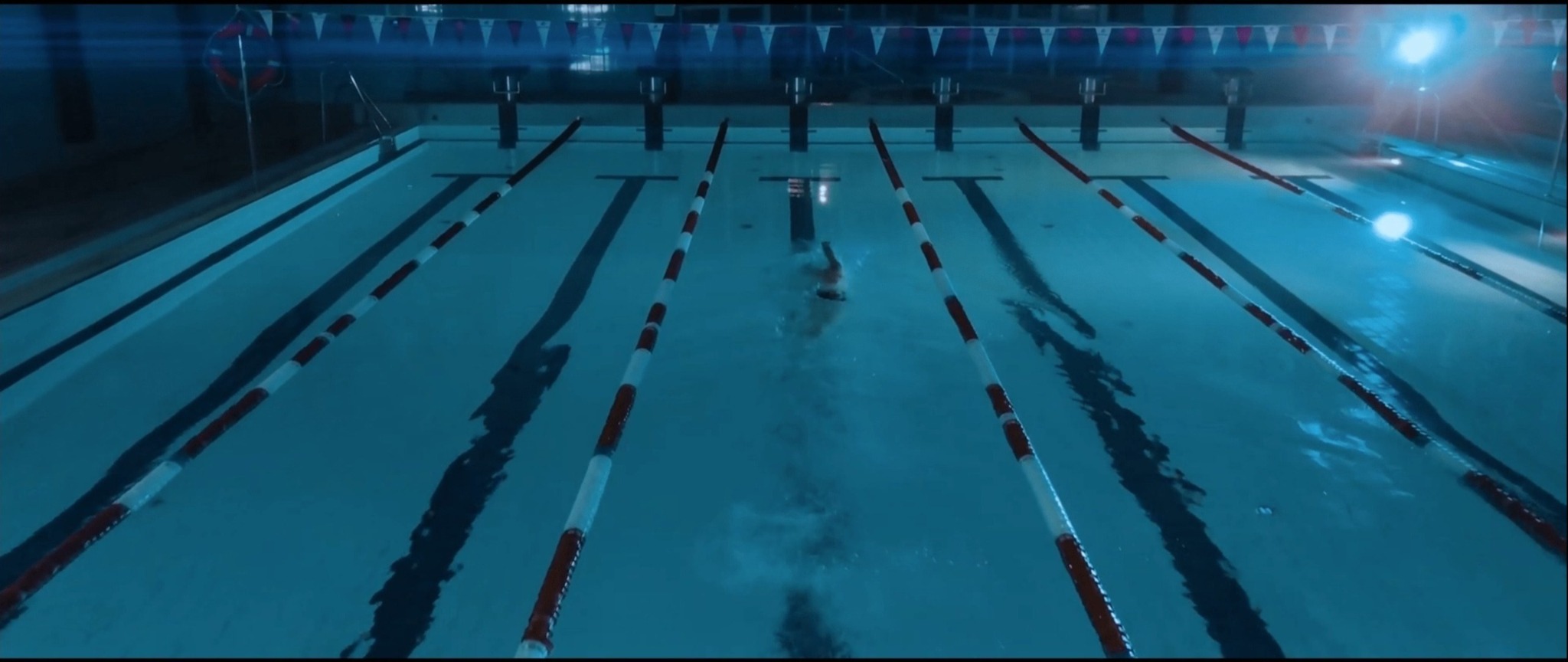 Kryty basen z torami pływackimi, w jednym z torów pływa osoba stylem dowolnym, oświetlenie górne.
