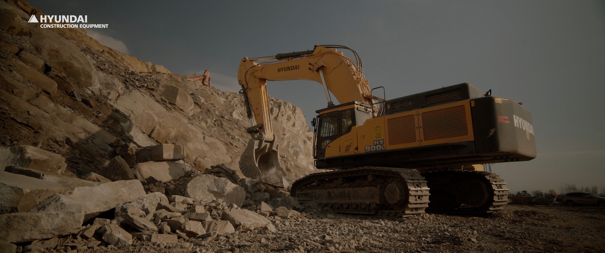 Żółta koparka Hyundai HX900L w trakcie pracy w kamieniołomie, z widocznymi blokami skalnymi i inną koparką w tle.