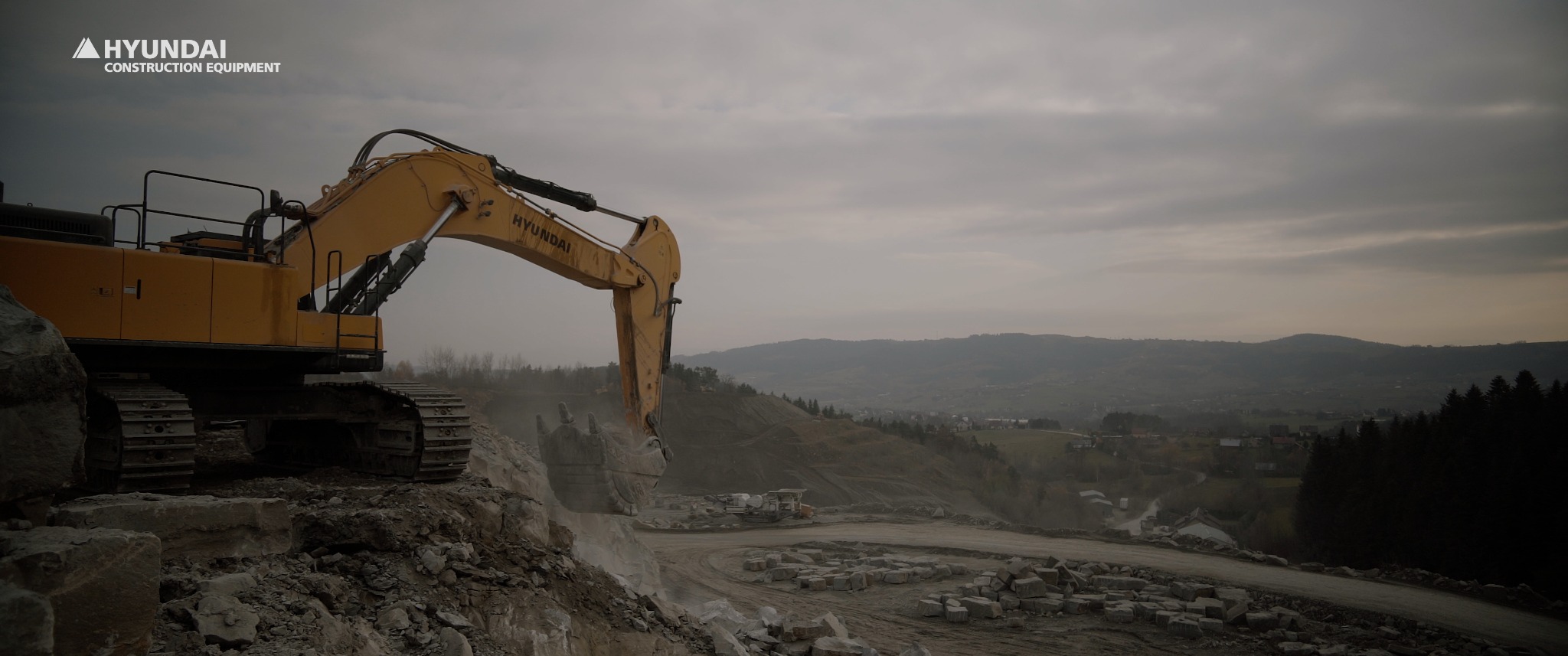 Żółta koparka Hyundai nabiera urobek skalny na tle pochmurnego krajobrazu zalesionych wzgórz.