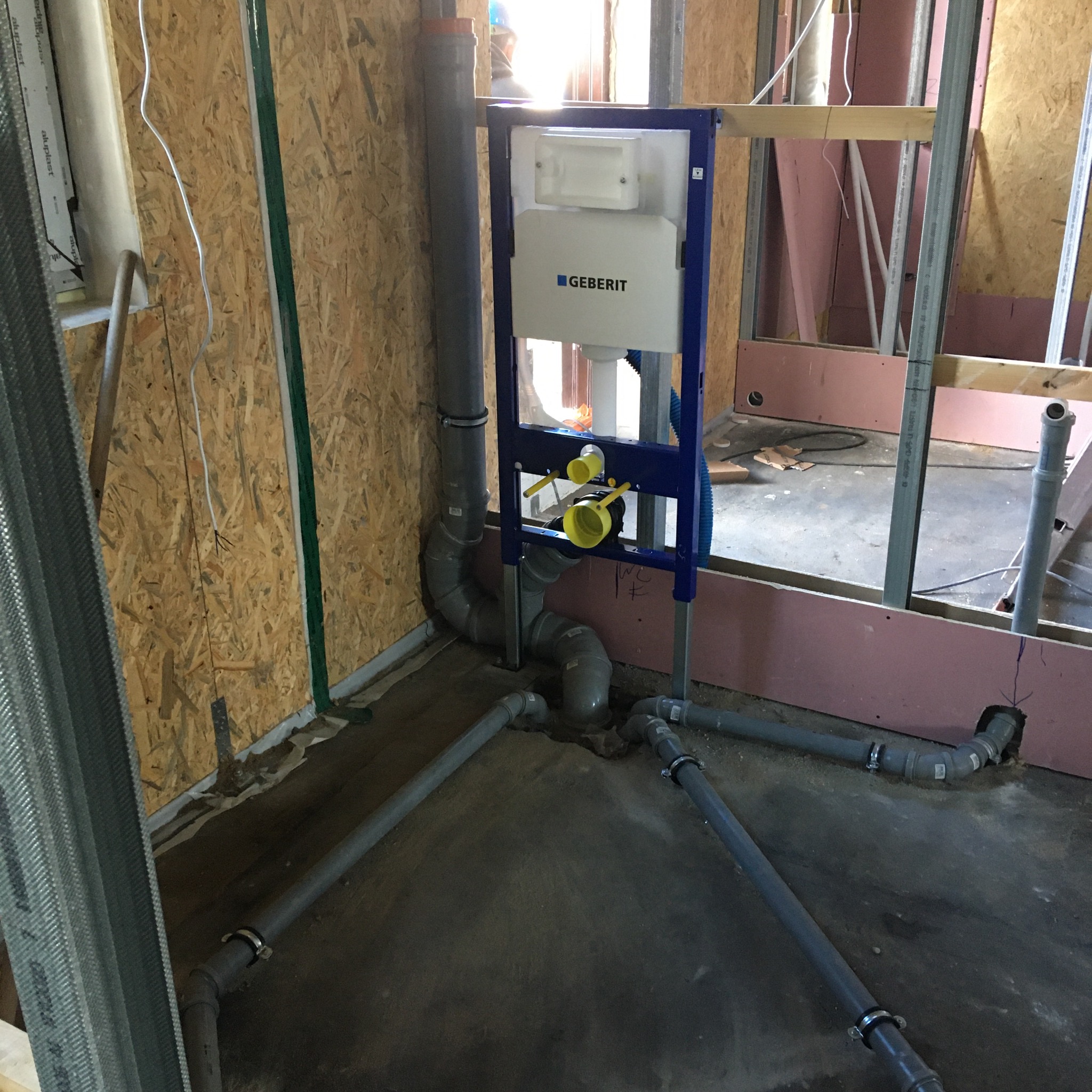 Surowa instalacja podtynkowa WC Geberit w trakcie montażu, widoczne szare rury kanalizacyjne i rama stelaża na tle ścian z płyty OSB i różowej wełny mineralnej.