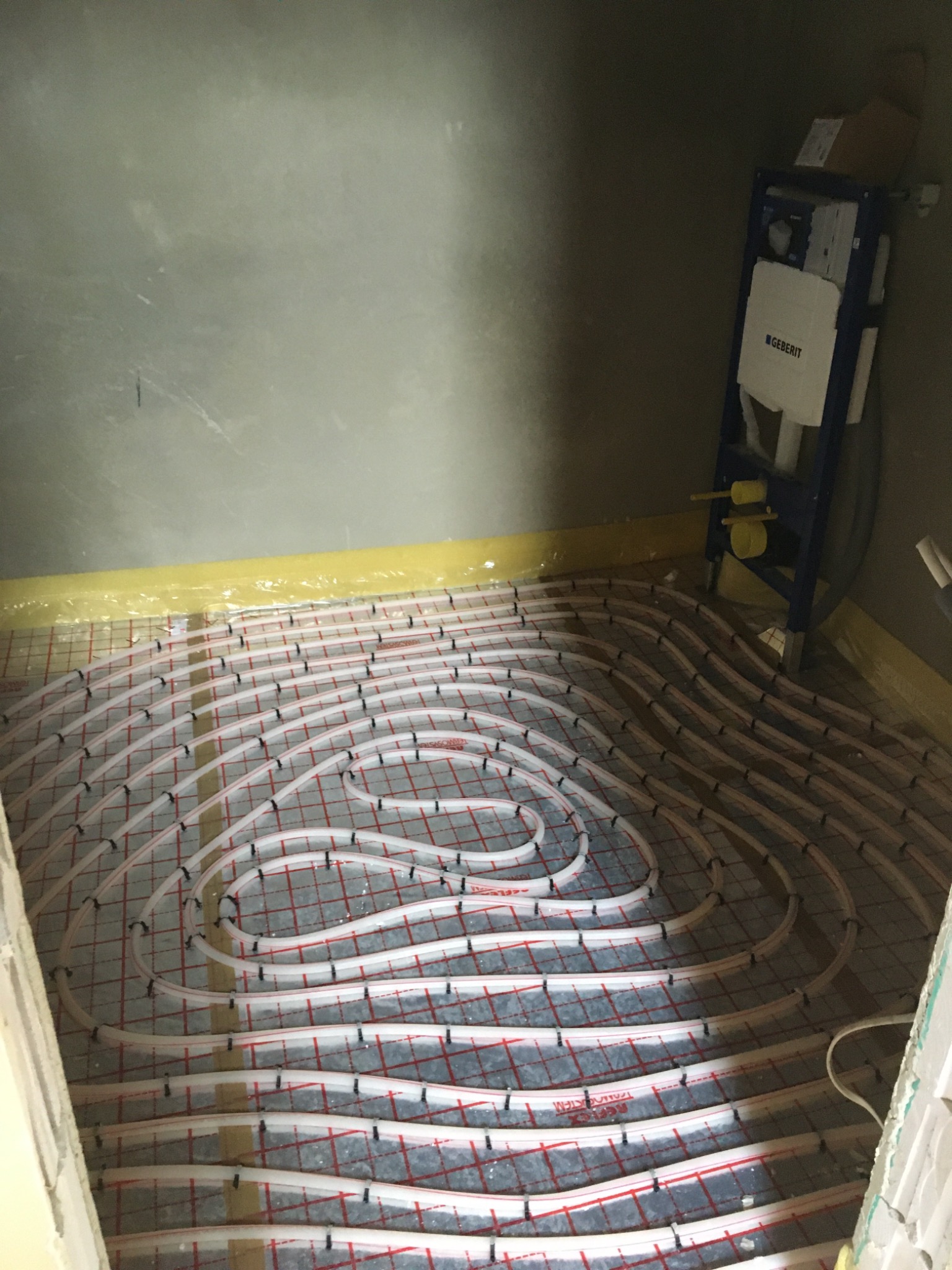 Rozłożone rury ogrzewania podłogowego na siatce z folią ochronną przy ścianach, widoczny stelaż podtynkowy Geberit.