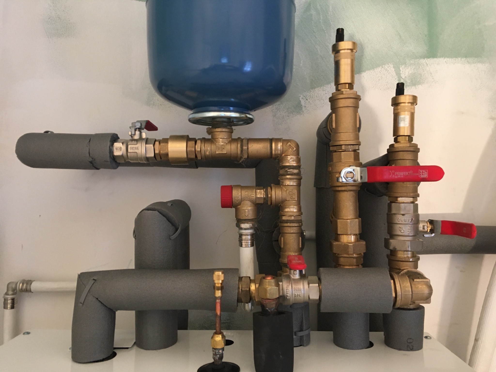 Fragment instalacji grzewczej z niebieskim zbiornikiem wyrównawczym, zaworami z czerwonymi rączkami i zaizolowanymi rurami w szarej otulinie, widok z bliska na połączenia i elementy instalacji.