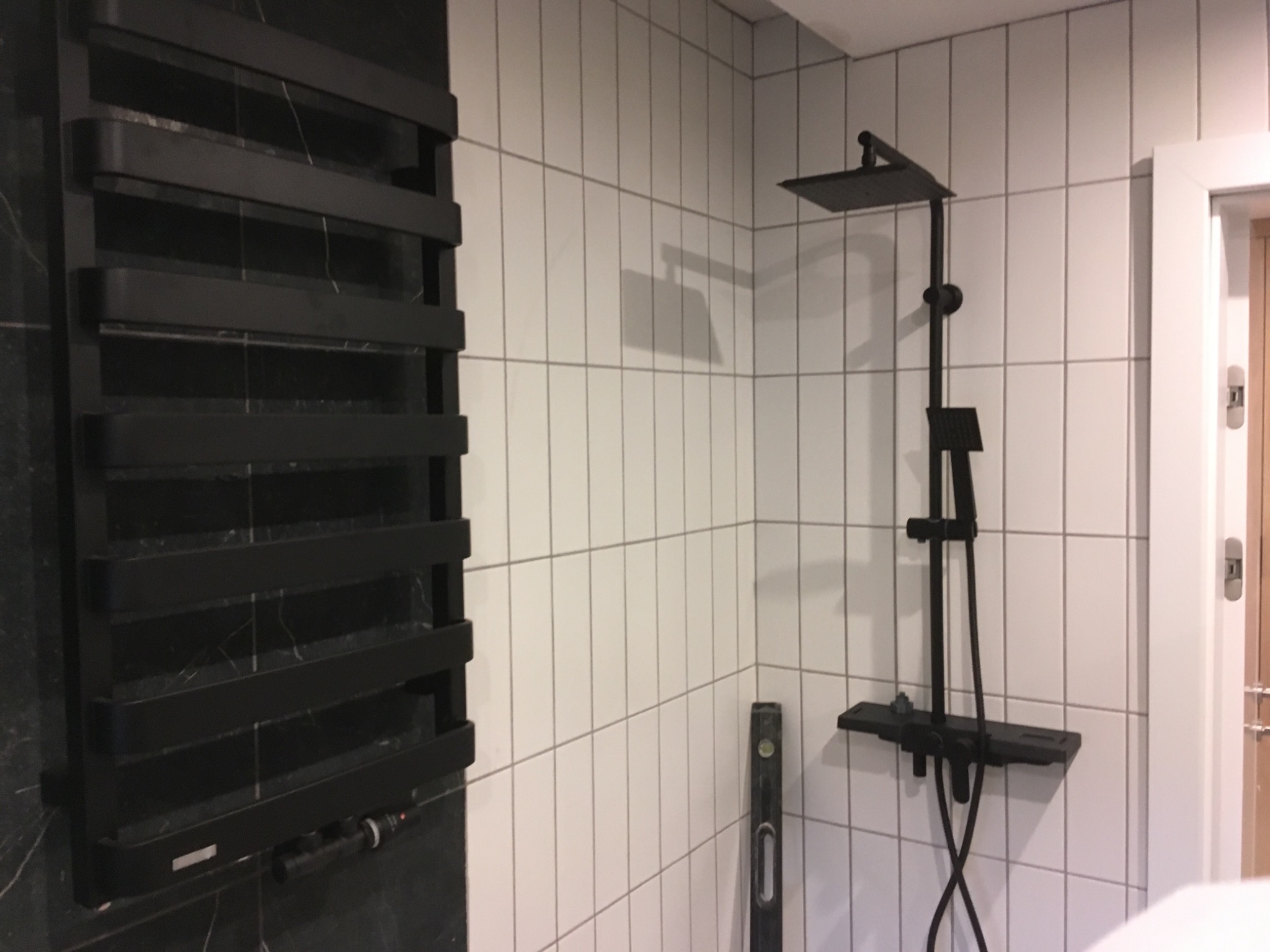 Nowoczesna łazienka z czarnym grzejnikiem drabinkowym na marmurowej ścianie i czarną armaturą prysznicową na białych, pionowych płytkach. Poziomica oparta o ścianę.