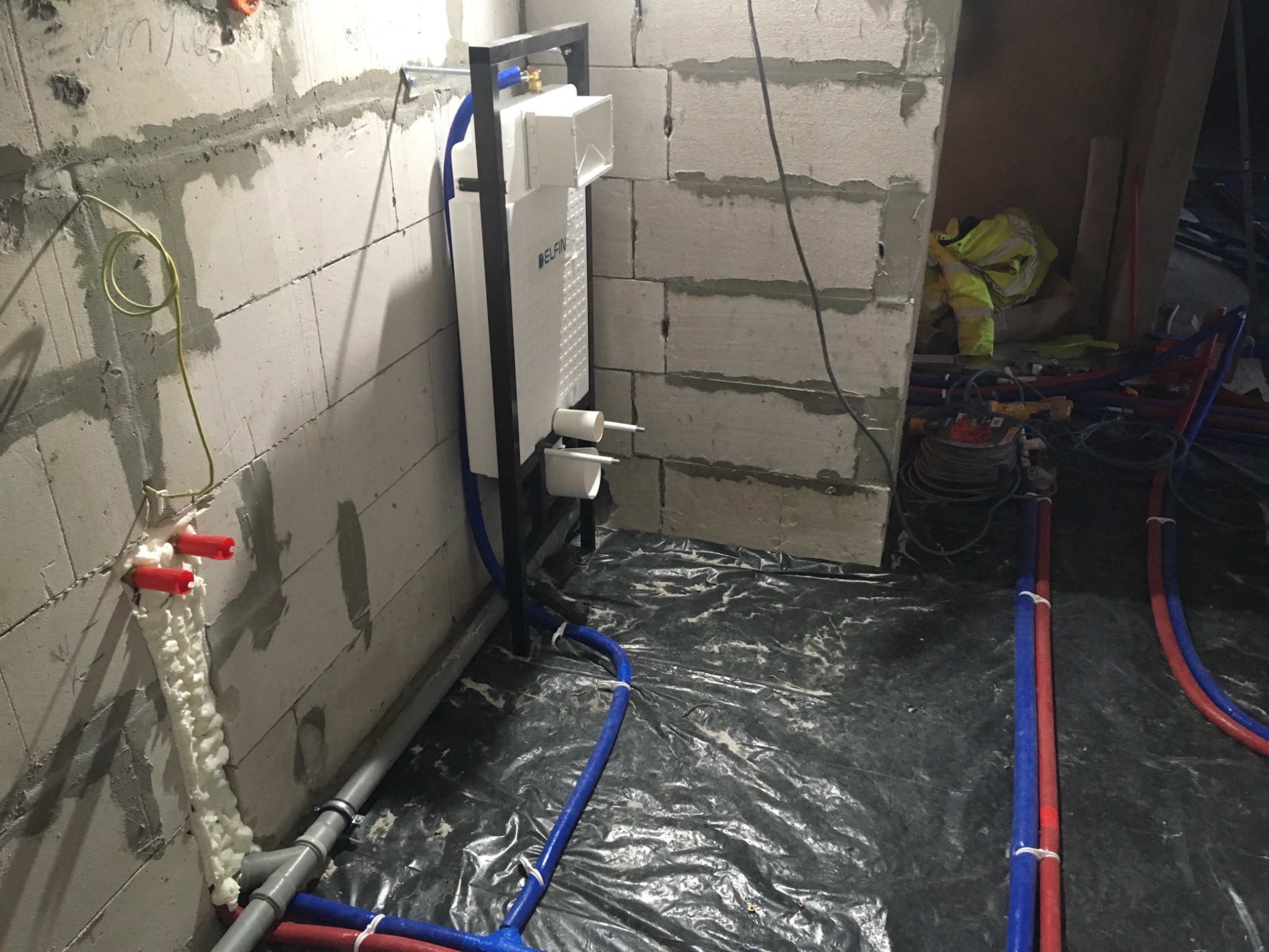 Montaż podtynkowego stelaża WC marki Delfin w surowym pomieszczeniu z widocznymi instalacjami wodno-kanalizacyjnymi, rurami PEX w kolorach czerwonym i niebieskim oraz zabezpieczonymi pianką...
