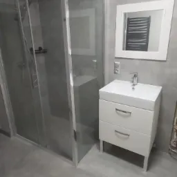 Nowoczesna łazienka z kabiną prysznicową z przezroczystego szkła, białą szafką pod umywalkę z dwiema szufladami i lustrem w białej ramie, odbijającym grzejnik drabinkowy.