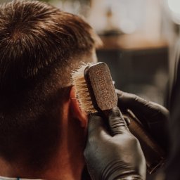 Cyrulicy na Koszutce | Barber Shop Katowice - Kark klienta podczas czesania szczotką z jasnym włosiem i drewnianą rączką z wygrawerowanym napisem, dłoń stylisty w czarnej rękawiczce, ciepłe oświetlenie.