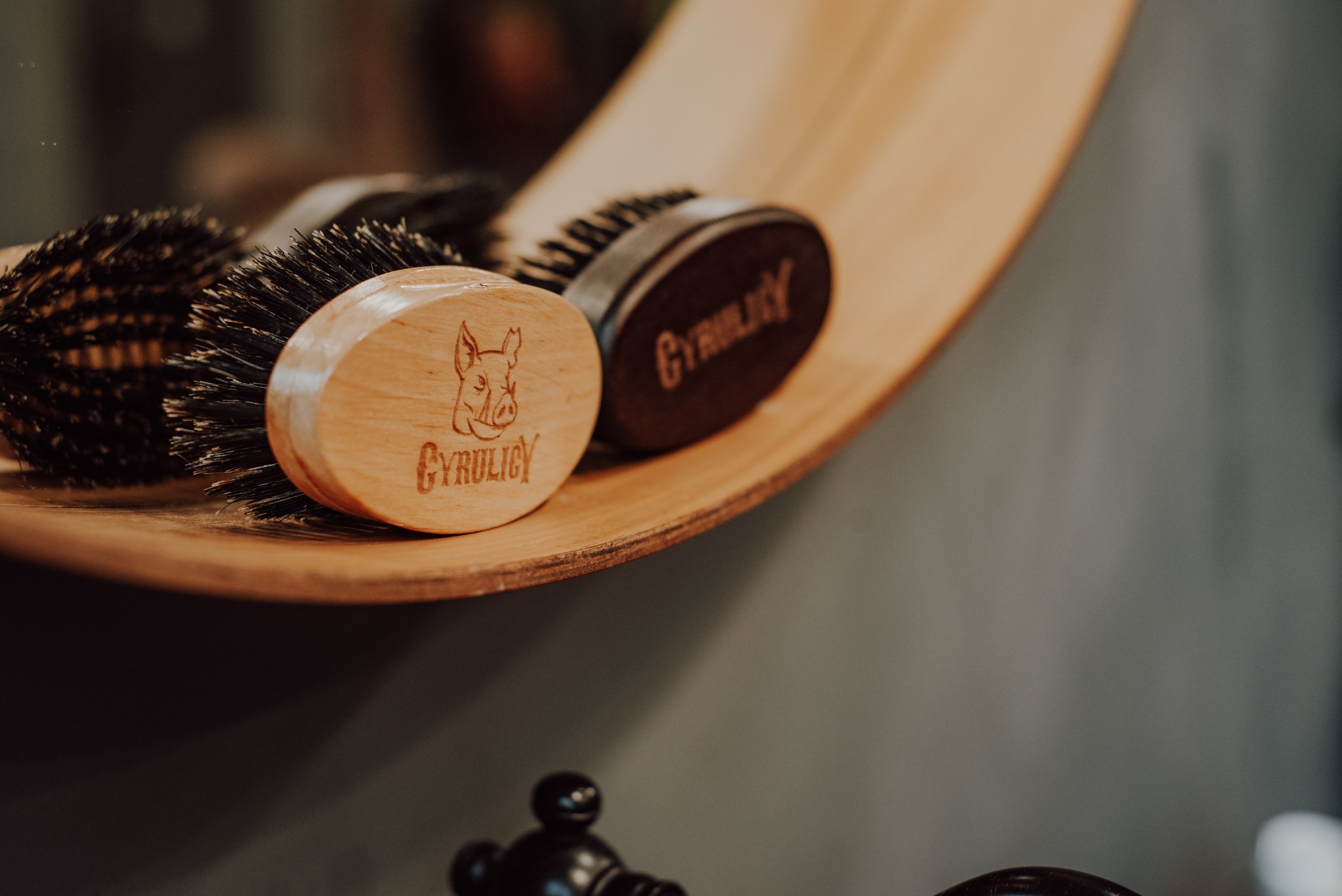 Dwie szczotki do brody z drewnianymi rączkami, jedna z wygrawerowanym wizerunkiem dzika, leżą na drewnianej półce.