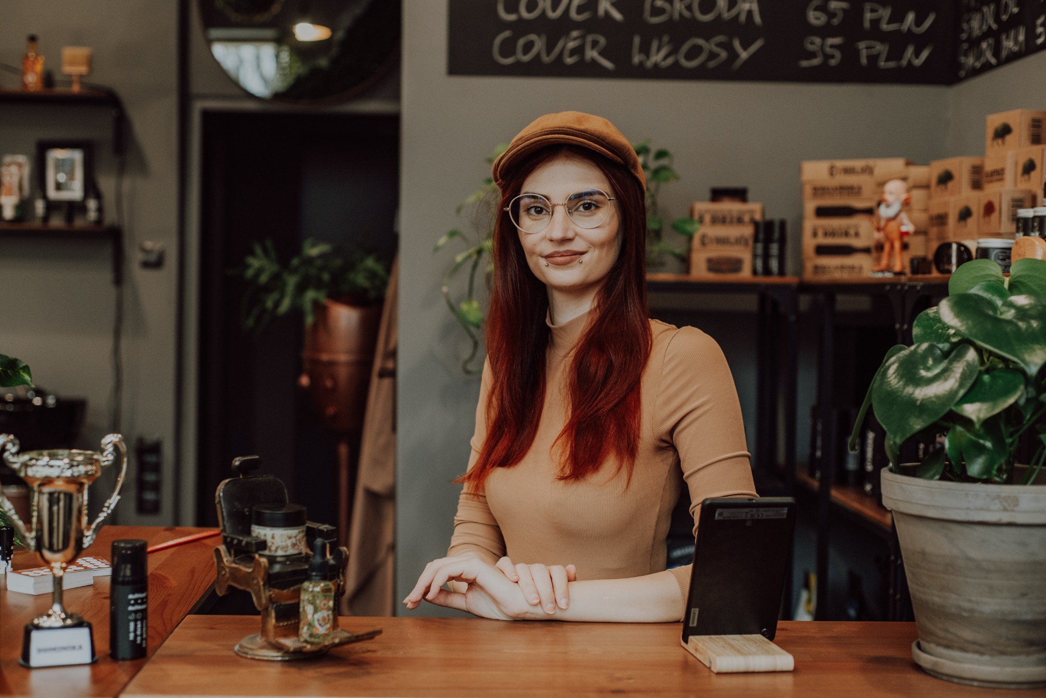 Uśmiechnięta kobieta w czapce kaszkietówce i okularach, siedząca za drewnianym blatem w salonie, na którym stoją akcesoria do pielęgnacji brody i włosów, w tle tablica z cennikiem usług.