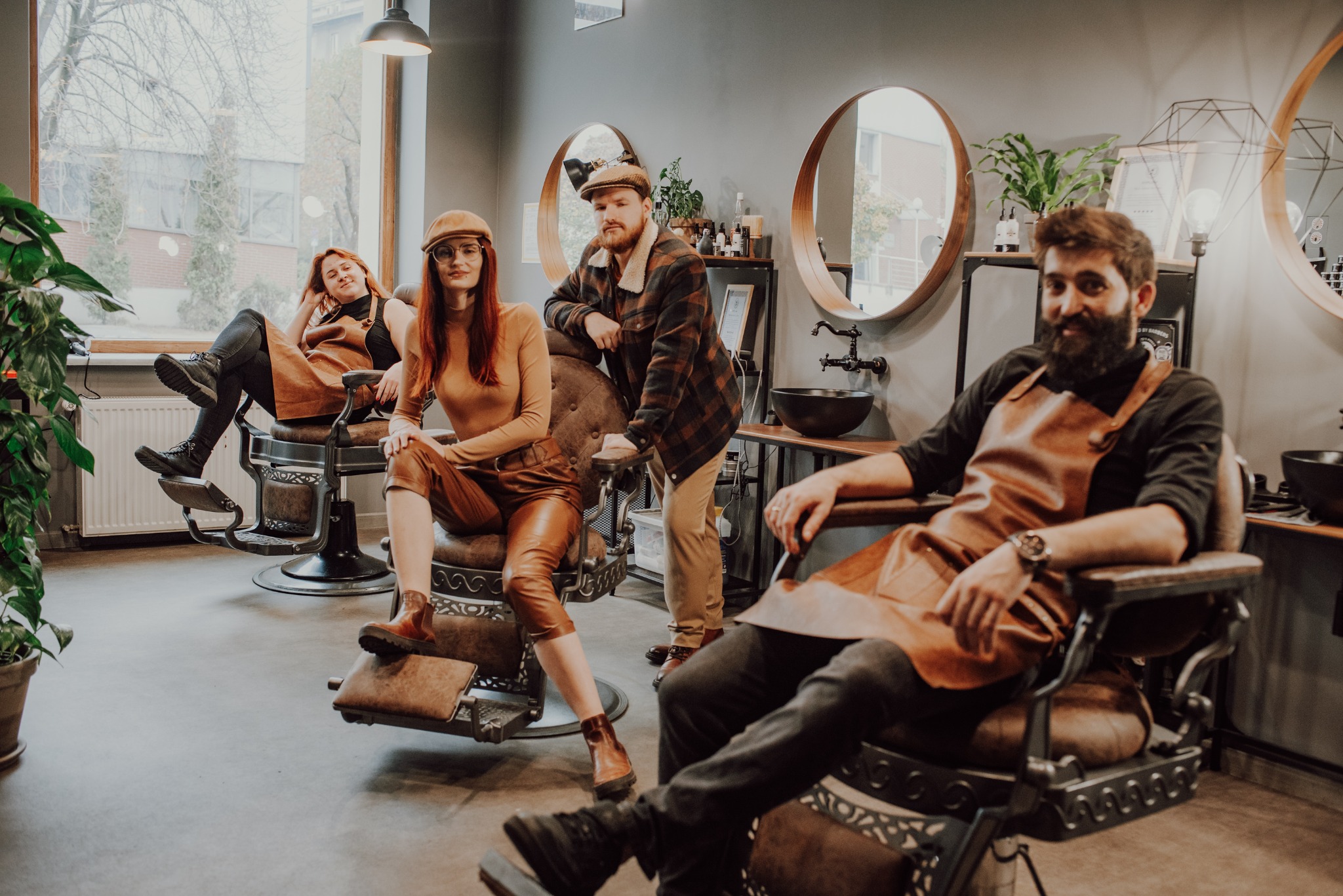Wnętrze salonu z czterema osobami w skórzanych fartuchach siedzącymi na fotelach fryzjerskich, w tle lustra i rośliny.
