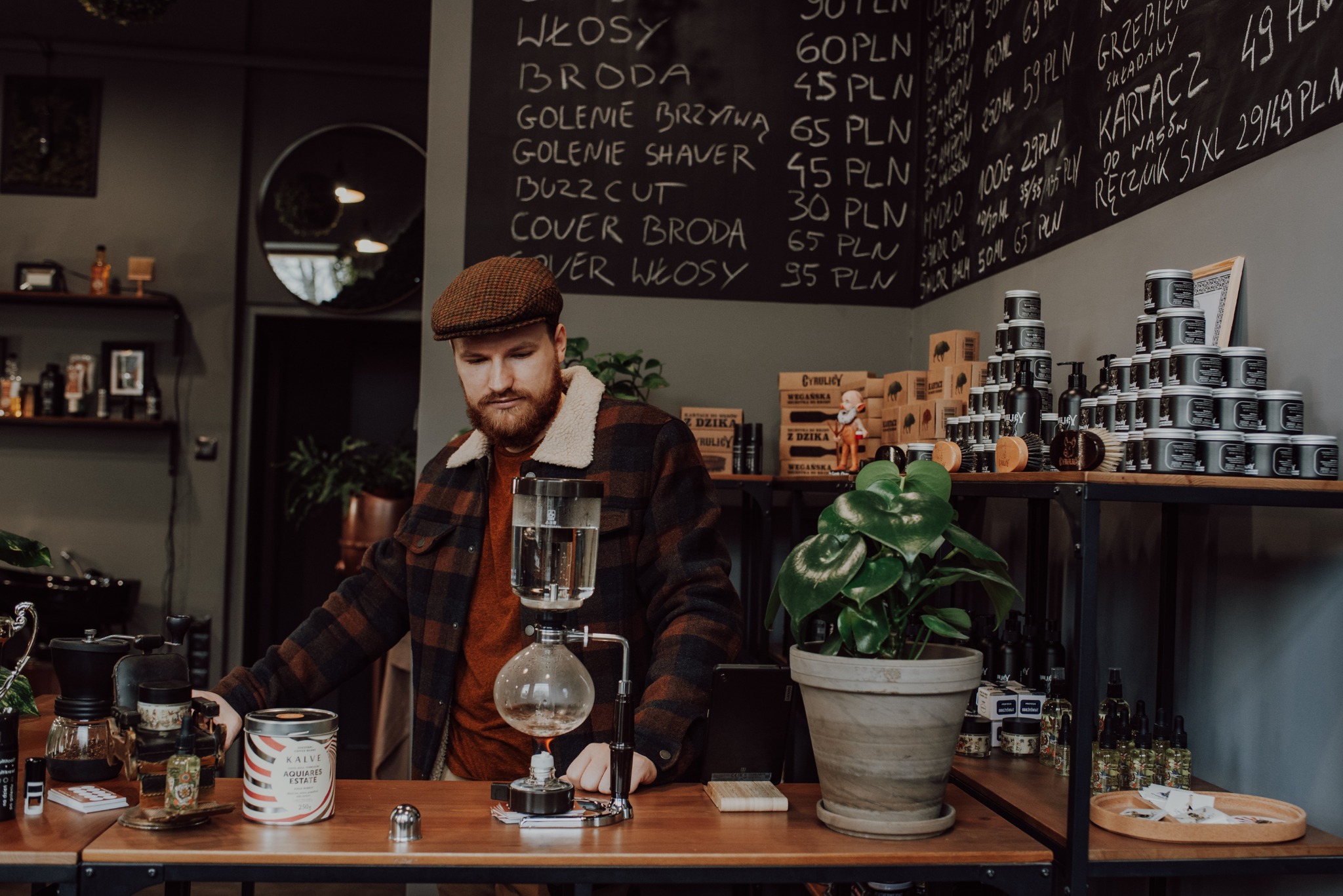Mężczyzna z brodą i w czapce przygotowuje napój za pomocą syfonu w eleganckim wnętrzu salonu, w tle tablica z cennikiem usług.