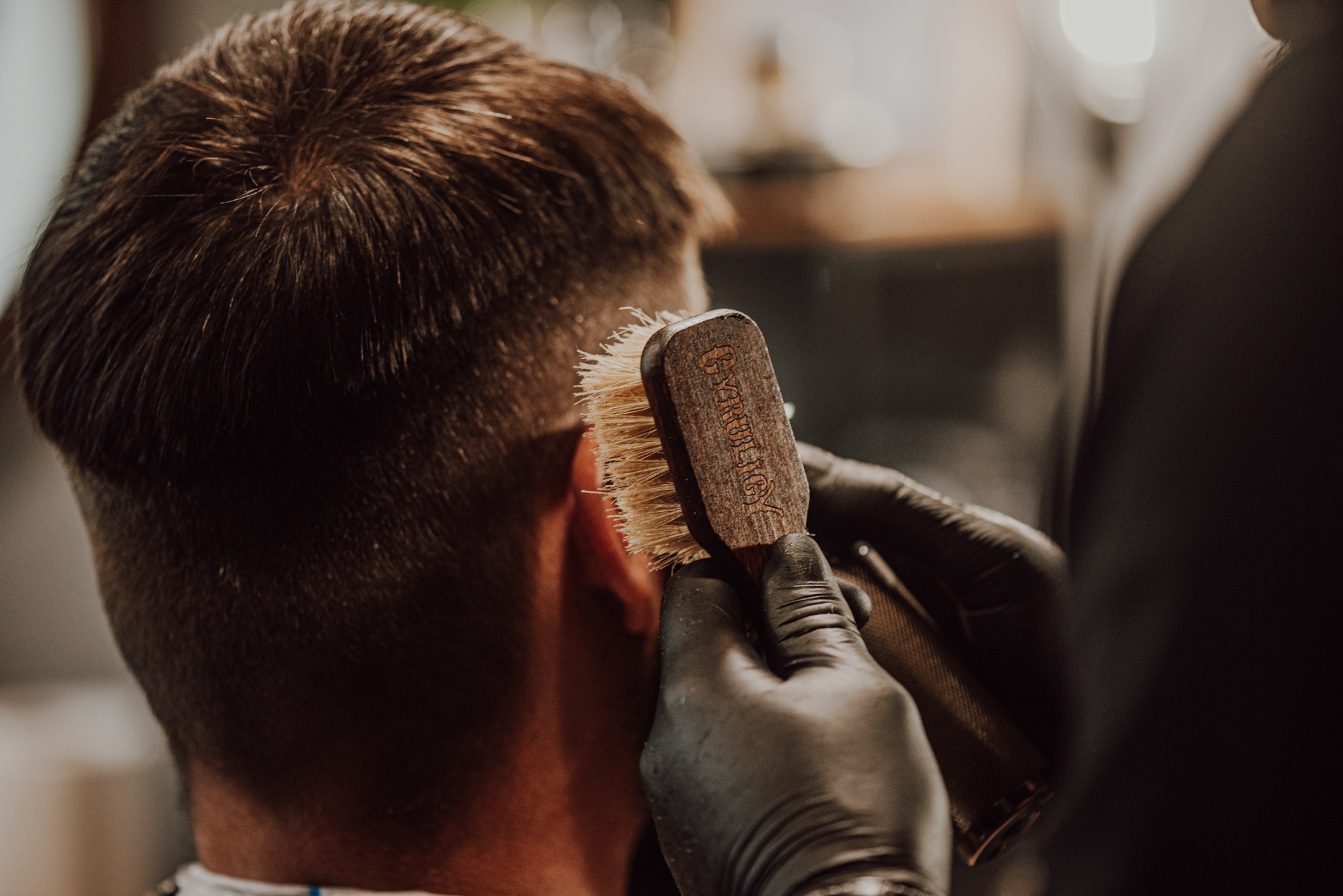 Kark klienta podczas czesania szczotką z jasnym włosiem i drewnianą rączką z wygrawerowanym napisem, dłoń stylisty w czarnej rękawiczce, ciepłe oświetlenie.