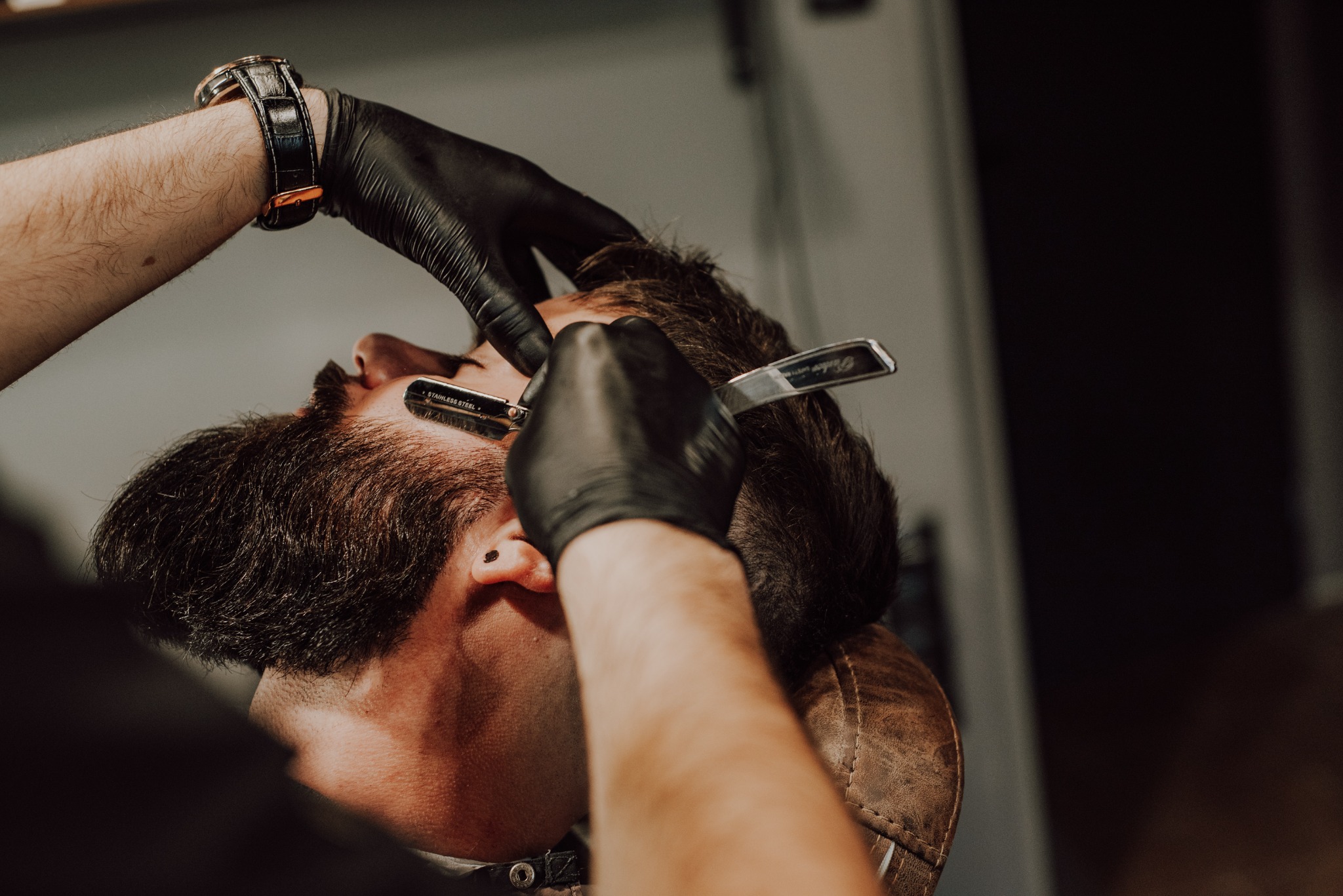 Fryzjer w czarnych rękawiczkach goli brodę klientowi brzytwą w salonie, widoczny zegarek na nadgarstku fryzjera.