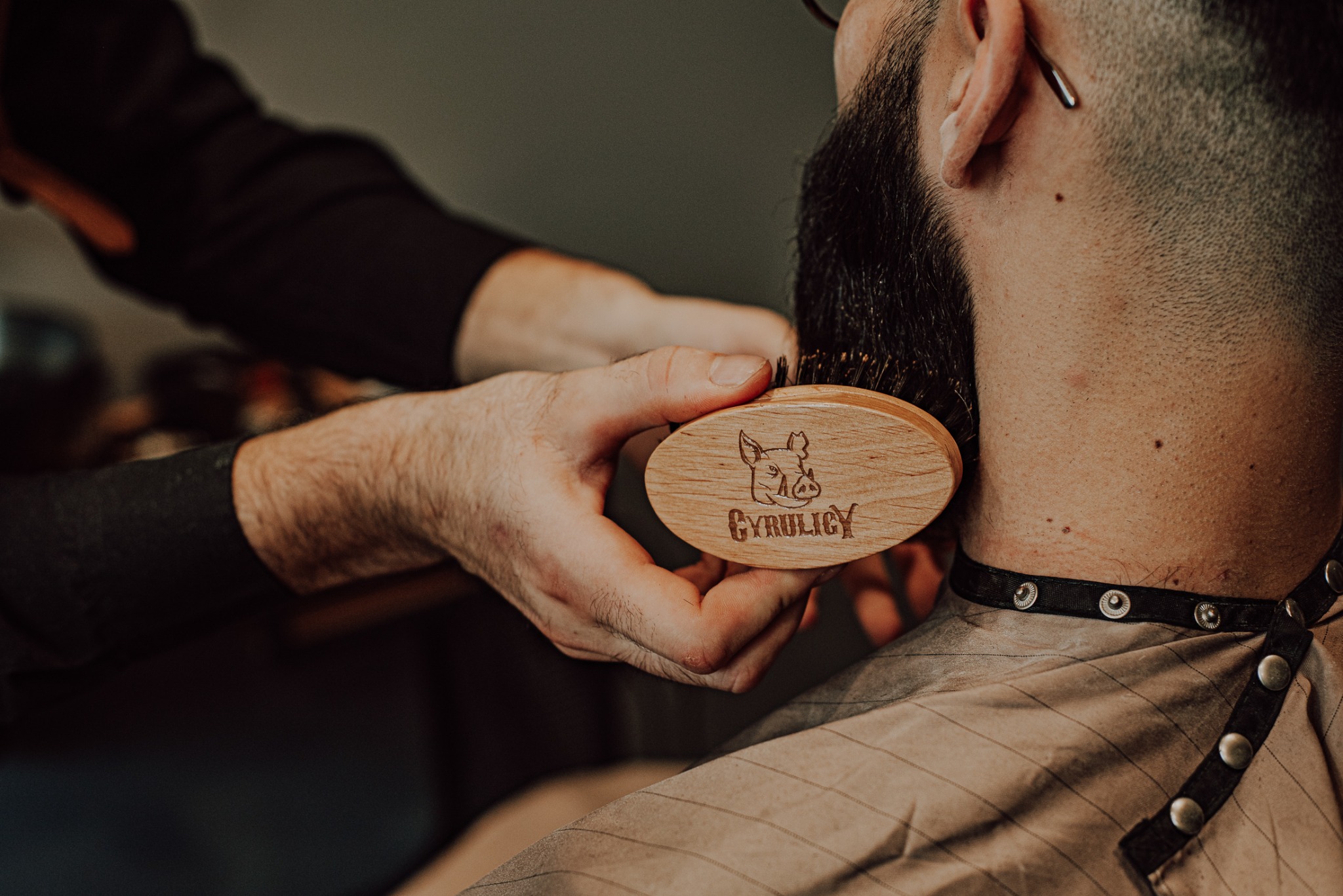 Stylizacja brody drewnianą szczotką z logo, widok z boku na brodę klienta w pelerynie z zapięciem na zatrzaski, dłoń stylisty w czarnym rękawie