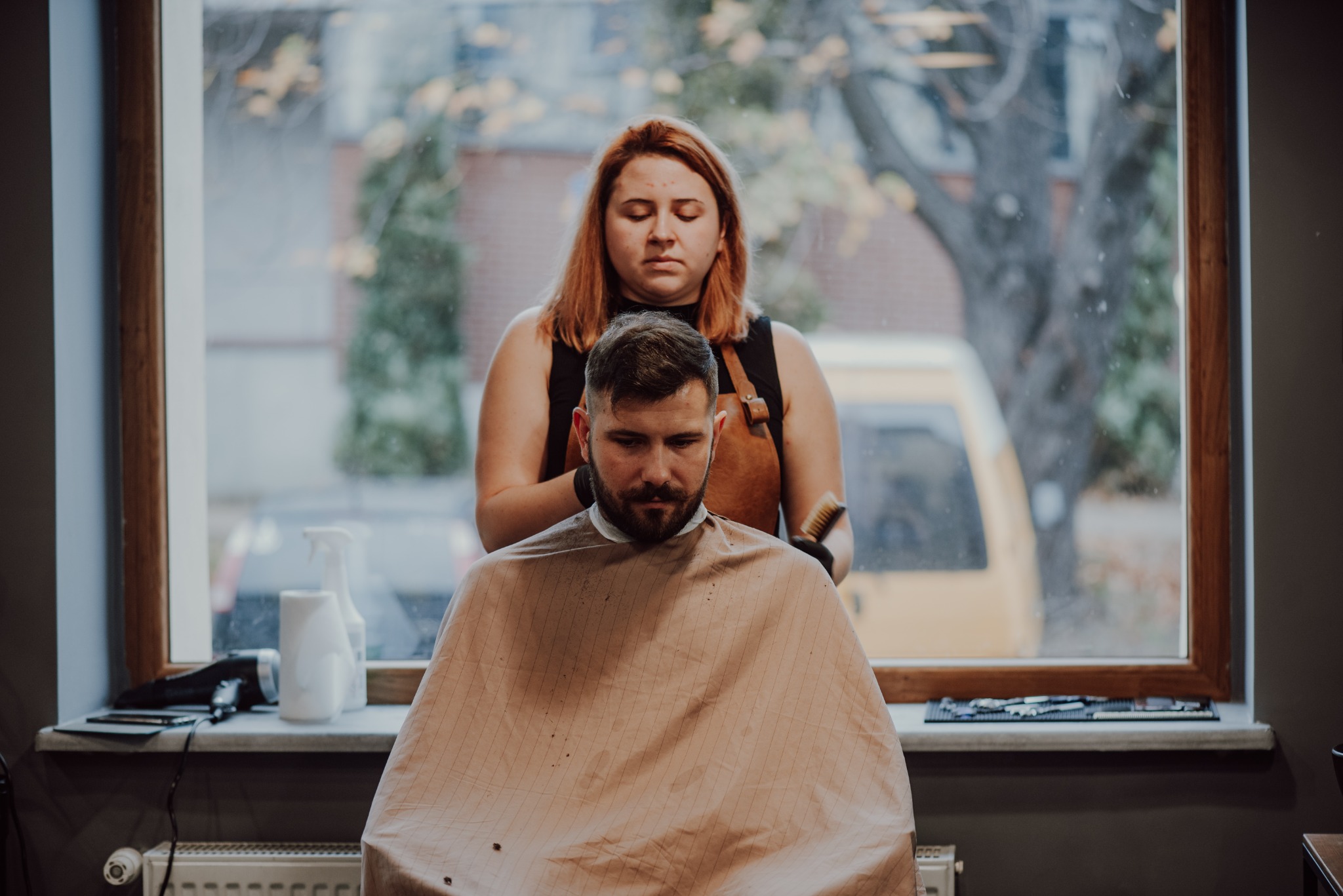 Klient z brodą siedzi w fotelu pod peleryną ochronną, a fryzjerka z rudymi włosami, w skórzanym fartuchu, trzyma szczotkę do włosów w trakcie strzyżenia, na tle okna z widokiem na ulicę.