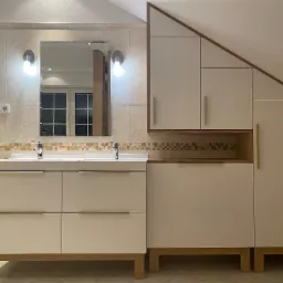 Biała szafka łazienkowa z dwiema umywalkami i lustrem, obok zabudowa pod skosem dachu z jasnymi frontami i drewnianymi akcentami, płytki w tle.