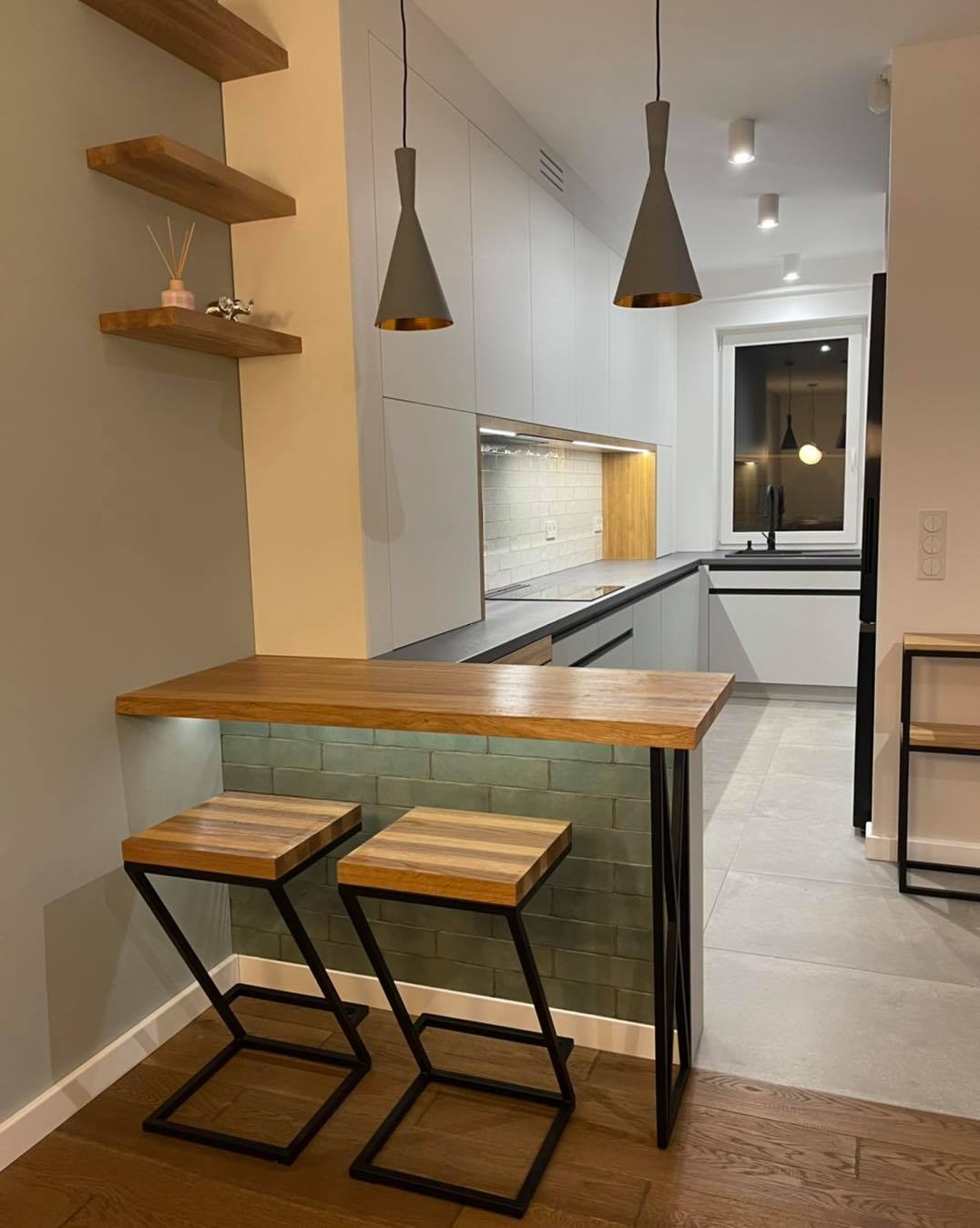 Nowoczesna kuchnia z białymi szafkami, drewnianym blatem i hokerami z metalowymi nogami, oświetlona wiszącymi lampami w kształcie stożków.