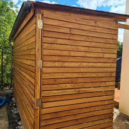 GoldenWood ANGELIKA DOLEGA - Drewniana konstrukcja, prawdopodobnie domek narzędziowy, wykonana z poziomo ułożonych desek w ciepłym, brązowym odcieniu, z widocznymi zawiasami i rynną, na tle zieleni i fragmentu budynku.