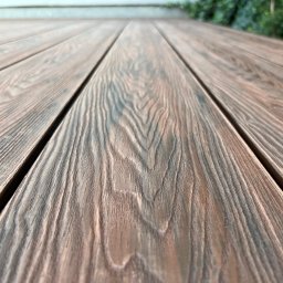 GoldenWood ANGELIKA DOLEGA - Deski tarasowe z wyraźnym usłojeniem, ułożone równolegle, perspektywa z dołu w górę, zieleń w tle. Układ desek tworzy geometryczny wzór.