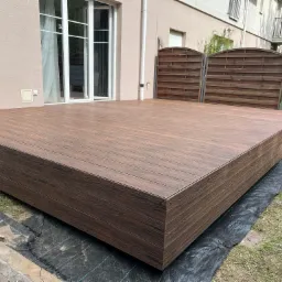 Nowoczesny taras w kolorze ciemnego drewna, z widoczną strukturą desek, tuż przy budynku z dużymi oknami i ogrodzeniem w tle. Widoczna czarna mata pod tarasem.