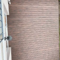 Kompozytowy taras w odcieniach brązu, ułożony przy ścianie budynku z oknem i lampą. Widok z góry, ukazujący strukturę desek i estetyczne wykończenie.