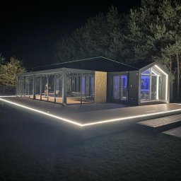 GoldenWood ANGELIKA DOLEGA - Nowoczesna zabudowa tarasowa z oświetleniem LED w Radomiu. Szklana konstrukcja połączona z drewnianym tarasem, oświetlona nocą, otoczona zielenią. Unikalny projekt.