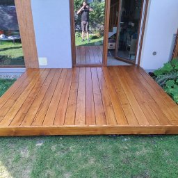 GoldenWood ANGELIKA DOLEGA - Nowo wybudowany taras drewniany z widocznymi słojami drewna, świeżo polakierowany, z trawnikiem w tle i odbiciem fotografa w szklanych drzwiach.