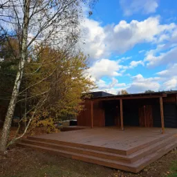 Nowoczesna, drewniana konstrukcja z podestem i zadaszeniem, otoczona drzewami w jesiennej scenerii, na tle błękitnego nieba z chmurami. Widoczny fragment zaparkowanego pojazdu z boku.