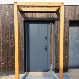 GoldenWood ANGELIKA DOLEGA - Szare drzwi wejściowe z drewnianą ramą i przeszklonym panelem bocznym, na tle ciemnej, drewnianej elewacji.