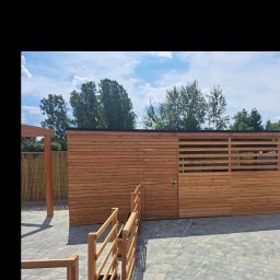 GoldenWood ANGELIKA DOLEGA - Drewniana konstrukcja z bramą i ażurowym panelem, prawdopodobnie garaż lub pomieszczenie gospodarcze, z widocznym fragmentem tarasu i ogrodzenia w tle, na tle błękitnego nieba z chmurami.