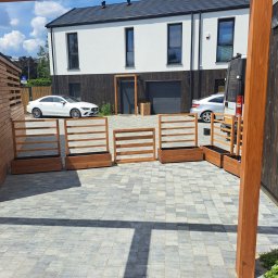 GoldenWood ANGELIKA DOLEGA - Drewniane donice z kratkami ustawione wzdłuż podjazdu z kostki brukowej, na tle nowoczesnego domu z białymi ścianami i ciemnymi elementami wykończeniowymi.