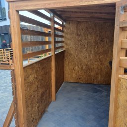 GoldenWood ANGELIKA DOLEGA - Wnętrze wiaty drewnianej z obiciem z płyty OSB, widok na zewnątrz przez ażurową ścianę, na zewnątrz pracownik podczas montażu drewnianej konstrukcji.