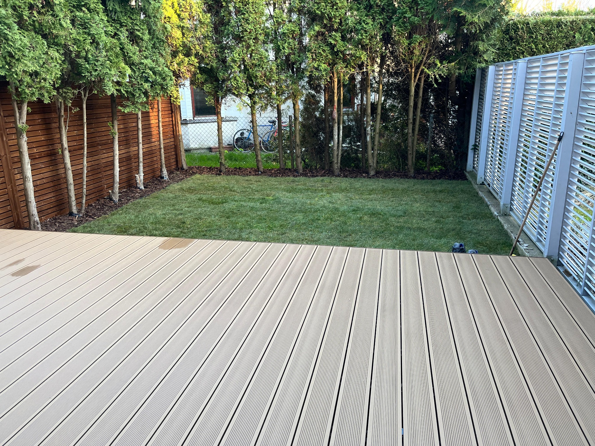 Jasny taras z desek kompozytowych przylegający do świeżo założonego trawnika, obok drewniany płot i rząd tuj, w tle rower oparty o siatkę.
