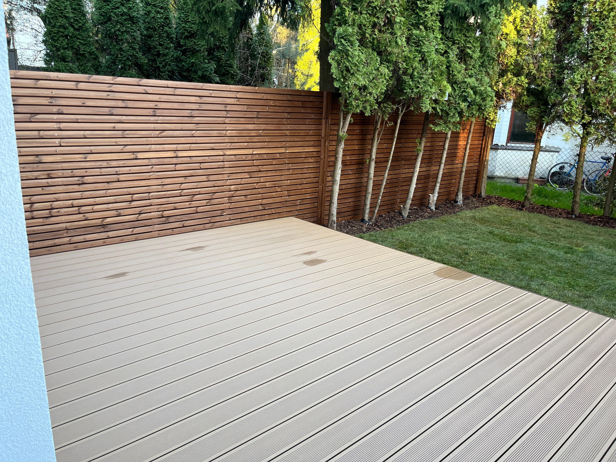 Jasny taras z desek kompozytowych sąsiadujący z drewnianym ogrodzeniem i trawnikiem, w tle widoczne tuje i rower oparty o siatkę ogrodzeniową.