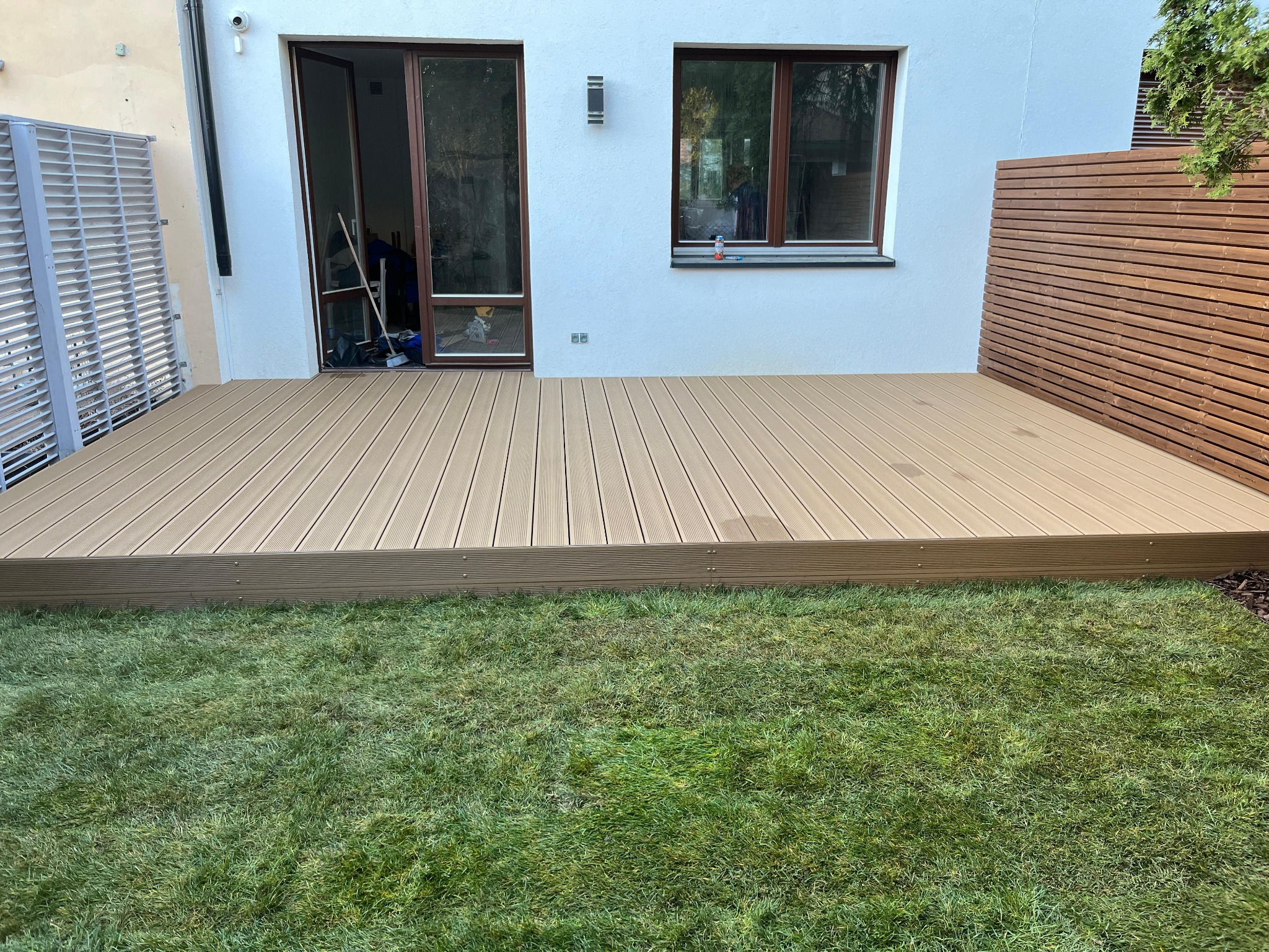 Nowoczesny taras z desek w kolorze jasnego drewna, przylegający do białego domu z brązowymi oknami, widok na trawnik i drewniane ogrodzenie.