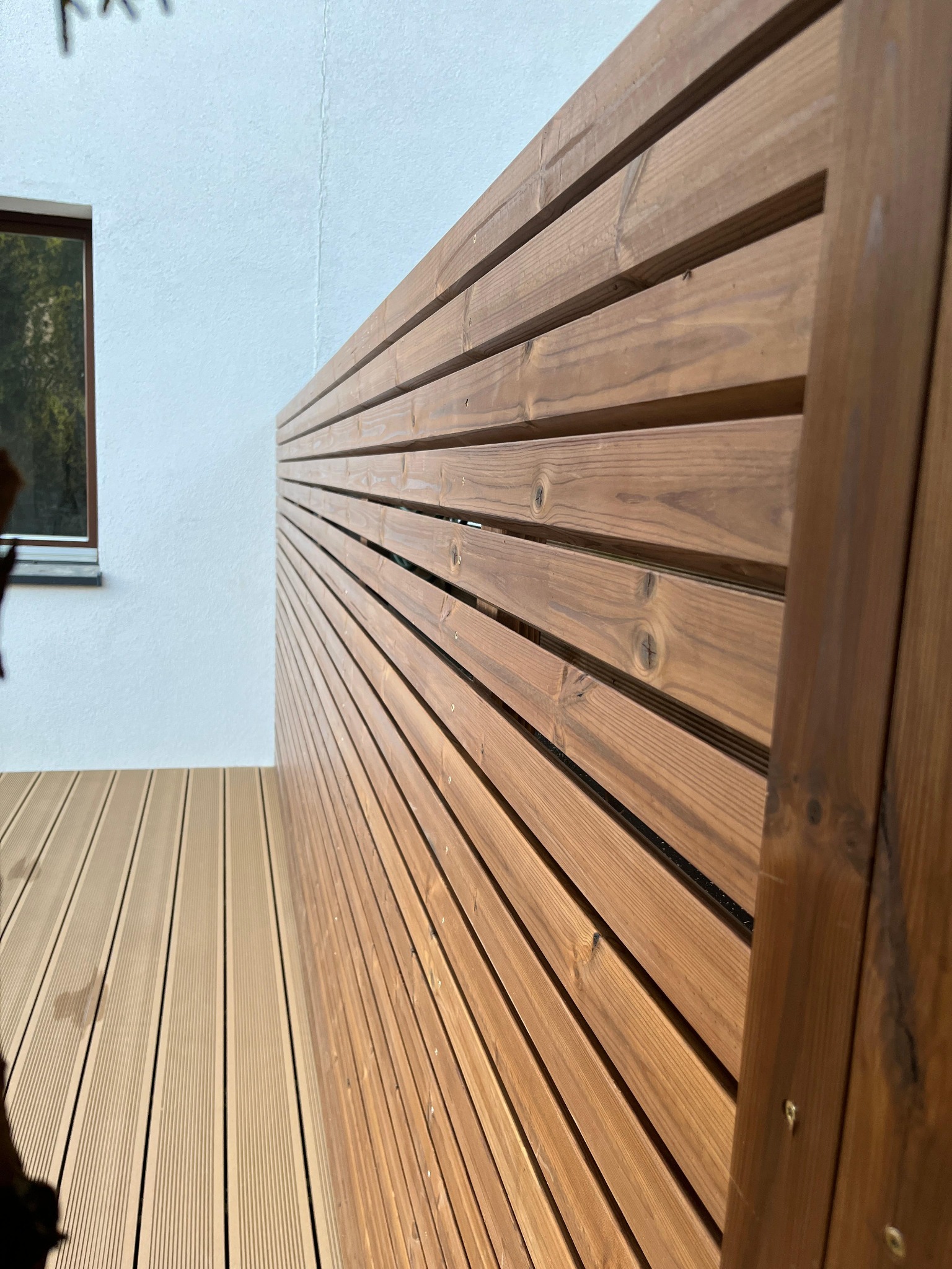 Drewniana, pozioma przesłona z desek, zamontowana na tarasie z desek kompozytowych, widok z boku, okno w tle.