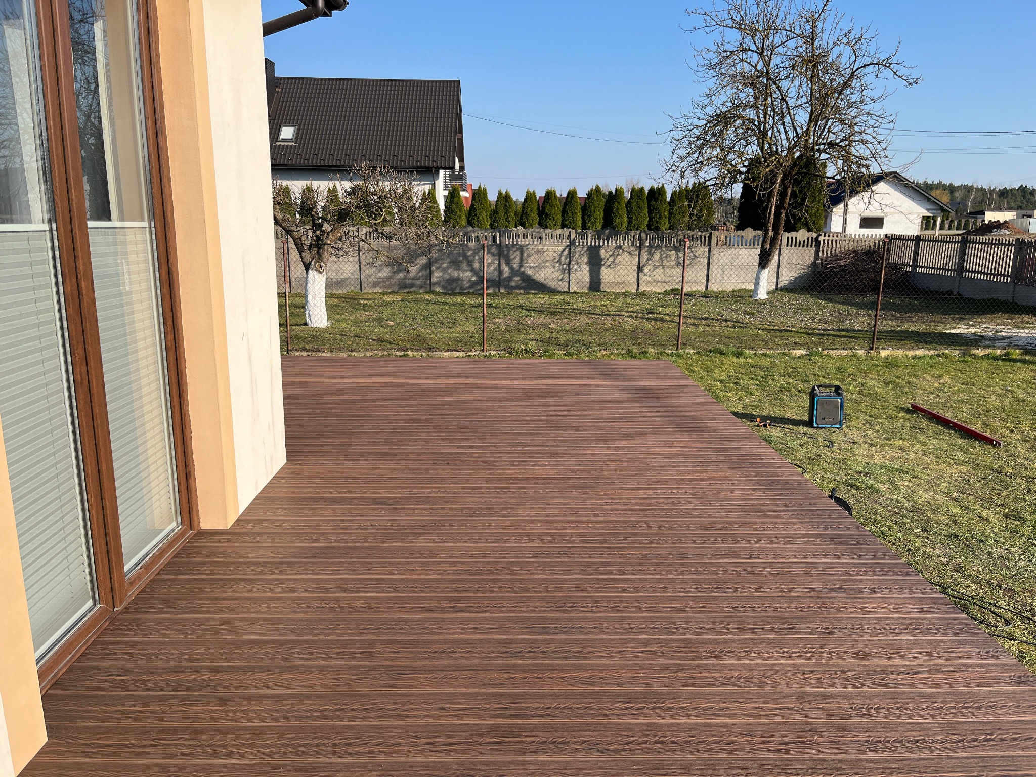 Brązowy taras z desek kompozytowych przylegający do domu, widok na ogród z trawnikiem, drzewami i ogrodzeniem.