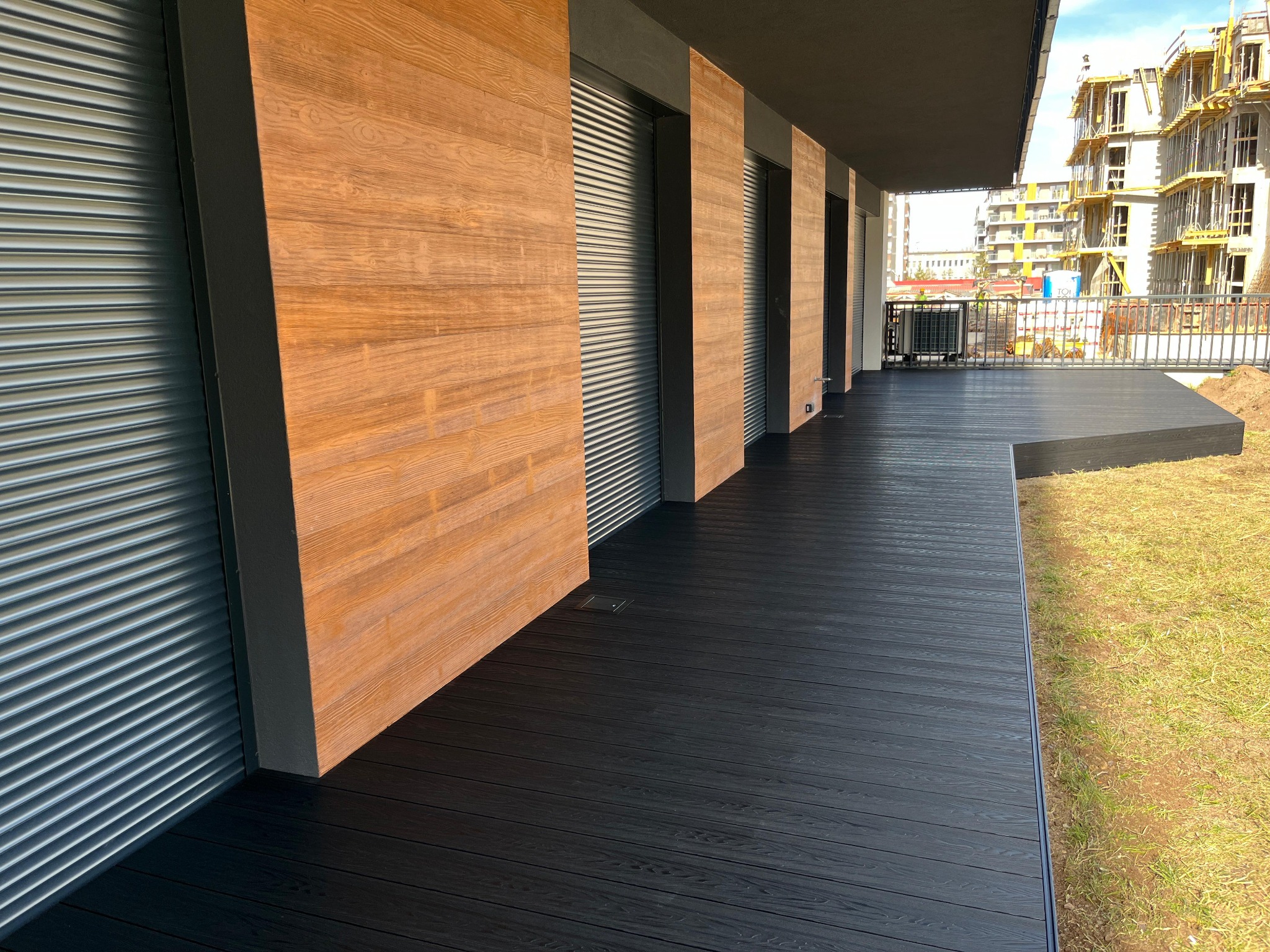Nowoczesny taras z ciemnych desek kompozytowych, przylegający do budynku z drewnianą elewacją i roletami zewnętrznymi, widok na plac budowy w tle.