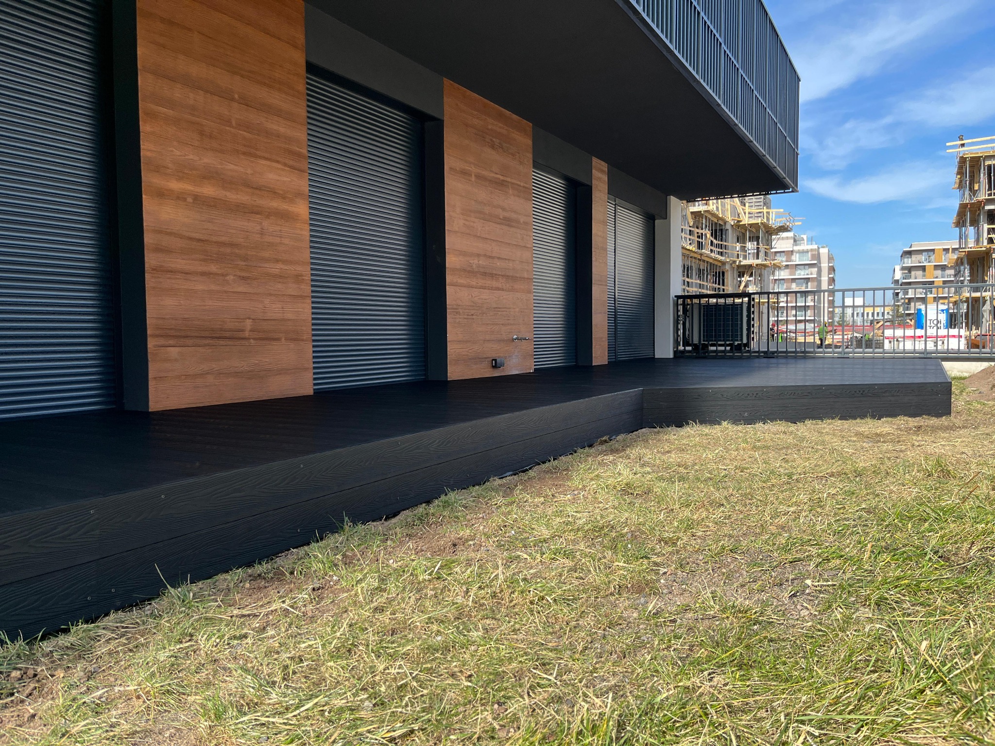 Nowoczesny budynek z czarnym tarasem i drewnianą elewacją, widok z poziomu trawnika, w tle budowa.