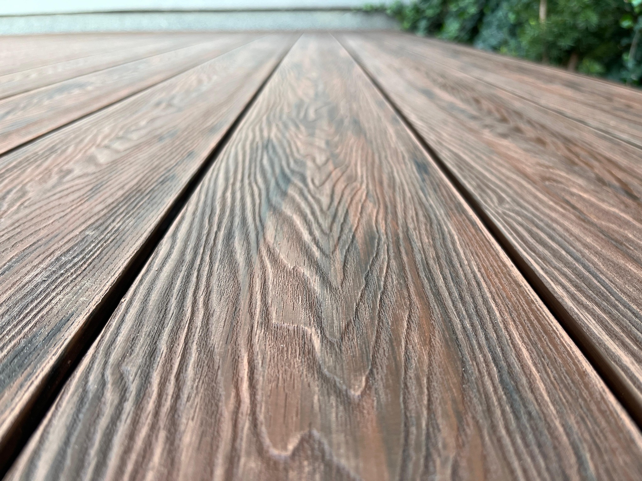 Deski tarasowe z wyraźnym usłojeniem, ułożone równolegle, perspektywa z dołu w górę, zieleń w tle. Układ desek tworzy geometryczny wzór.
