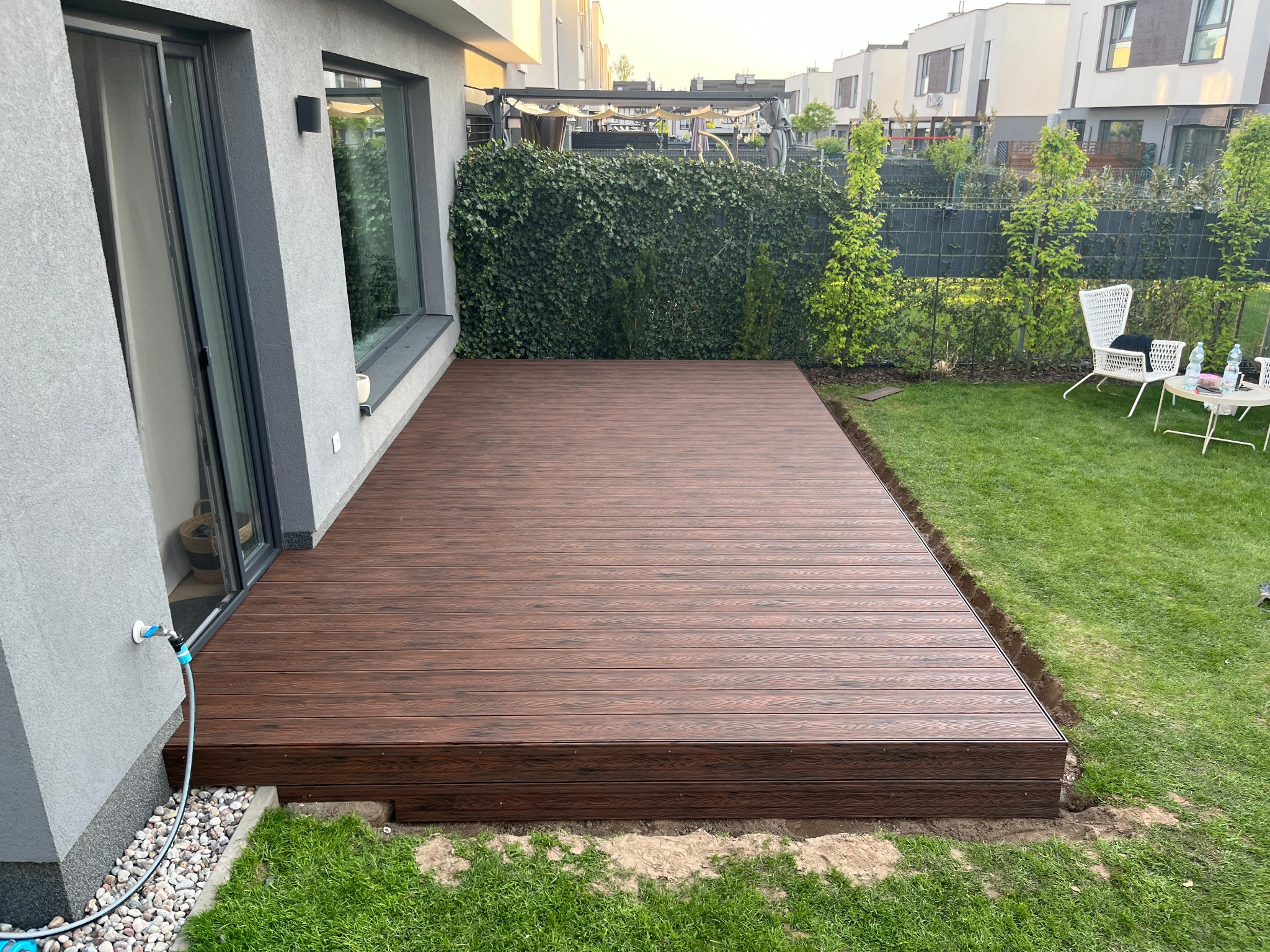 Nowoczesny taras z desek kompozytowych w kolorze ciemnego drewna, przylegający do domu z szarą elewacją. Widoczne trawnik i roślinność ogrodowa.