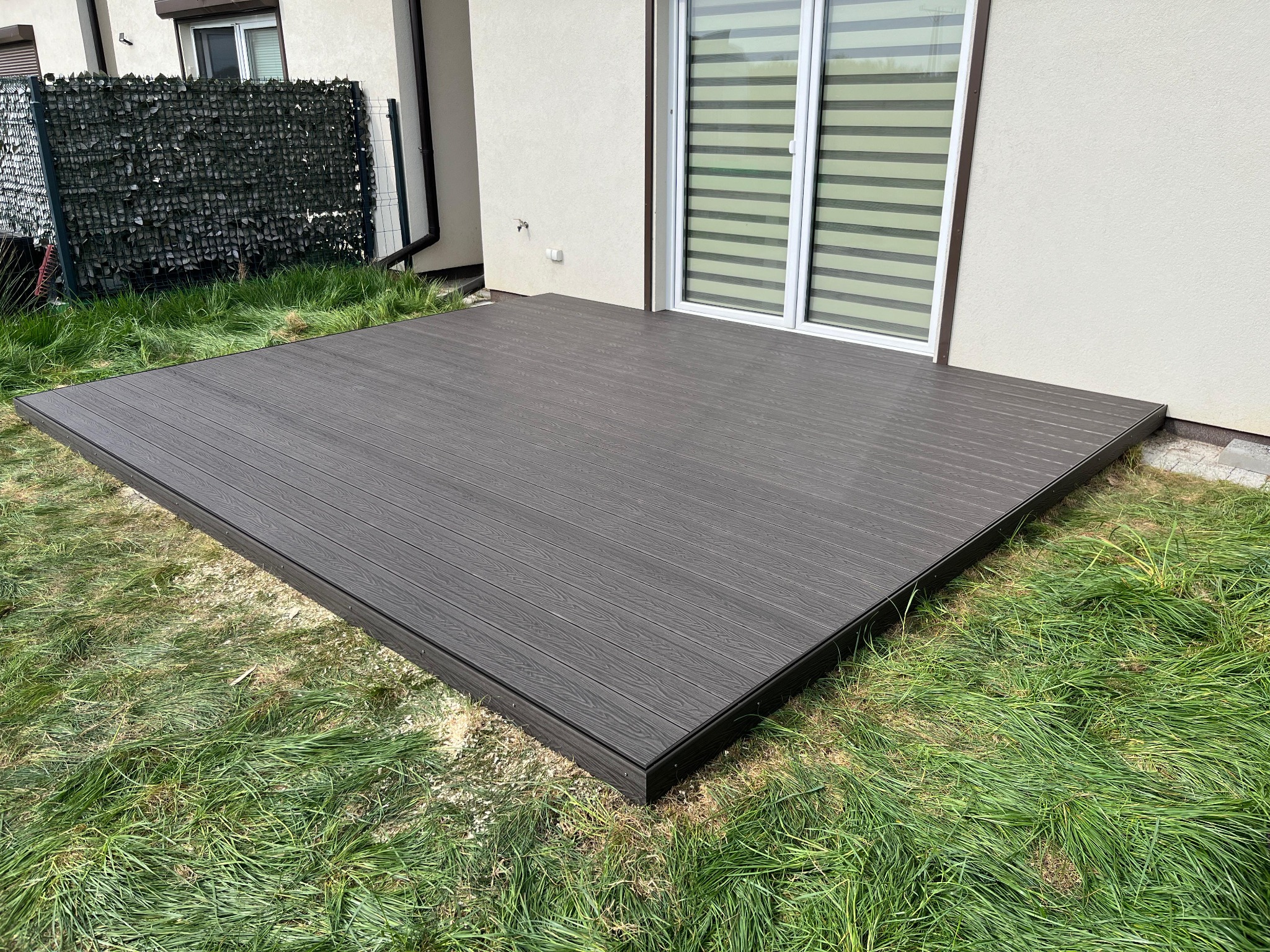 Nowoczesny, brązowy taras kompozytowy przylegający do budynku z oknami przesuwnymi, otoczony zieloną trawą. Widoczne detale łączeń desek i obrzeży tarasu.