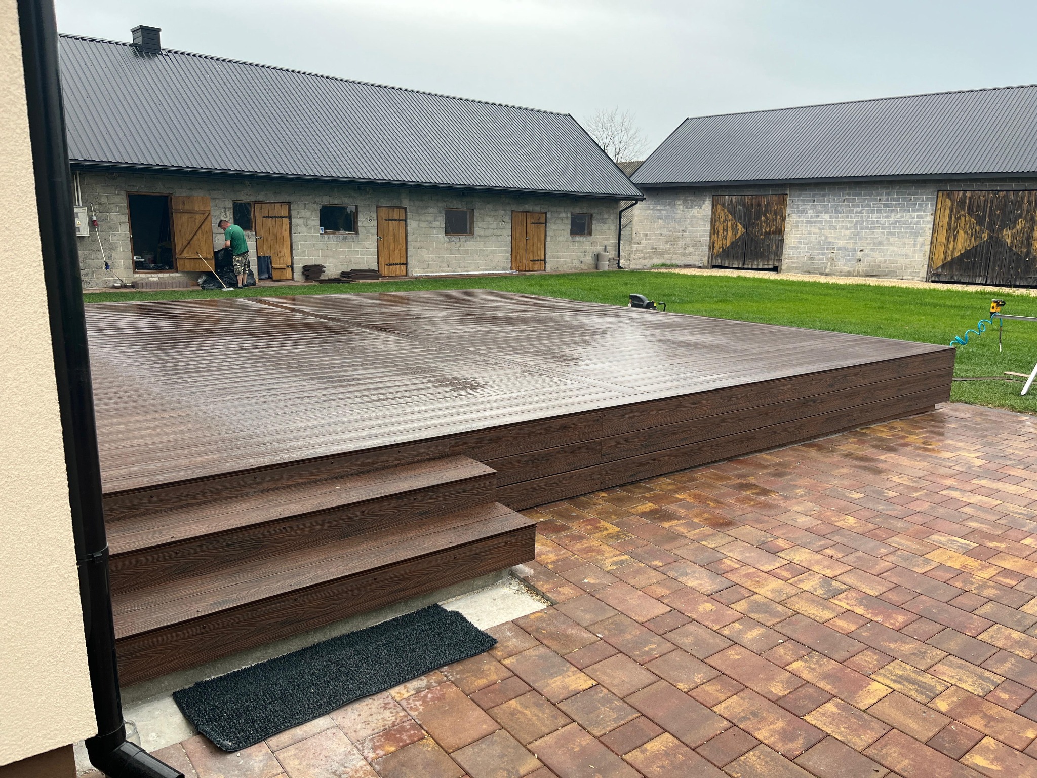 Nowoczesny taras z deski kompozytowej w odcieniach brązu, z niskimi schodami i kostką brukową w kolorach jesieni. W tle budynek gospodarczy z szarą blachodachówką.